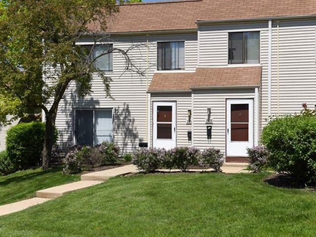 Two-story beige townhouse with a green lawn, front walkway, and two red doors.
