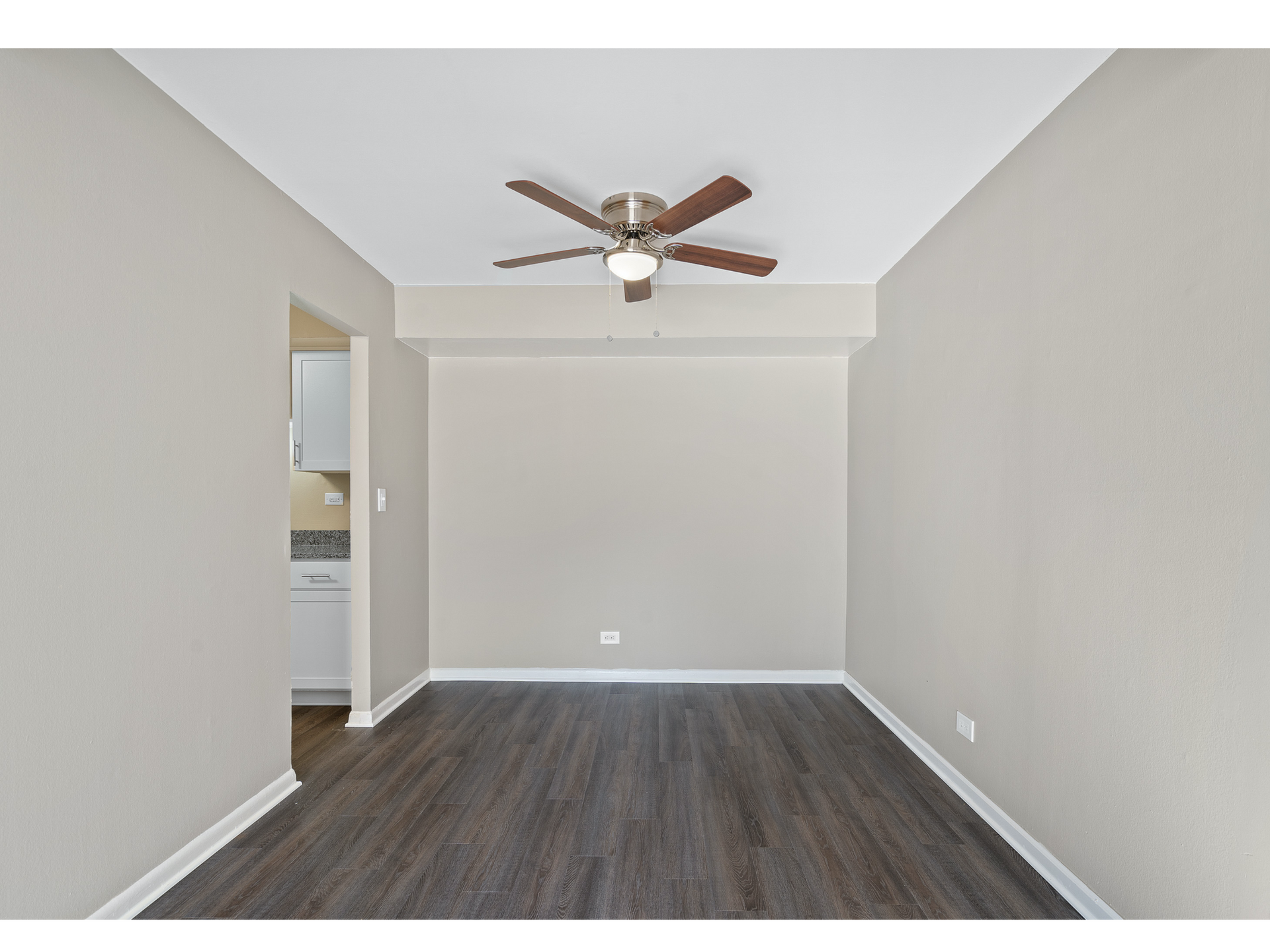 Empty beige room with dark wood flooring, ceiling fan, and doorway to a bathroom