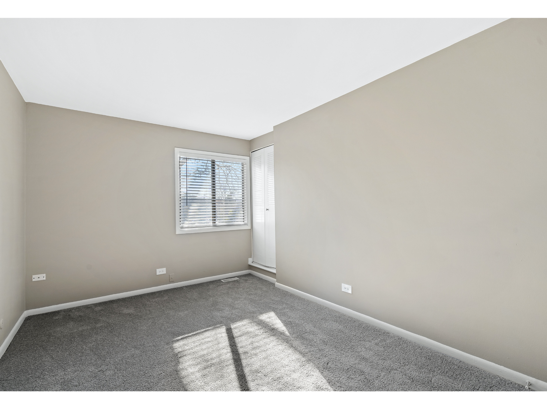 Empty beige bedroom with gray carpet, a window, and sunlight on the floor.