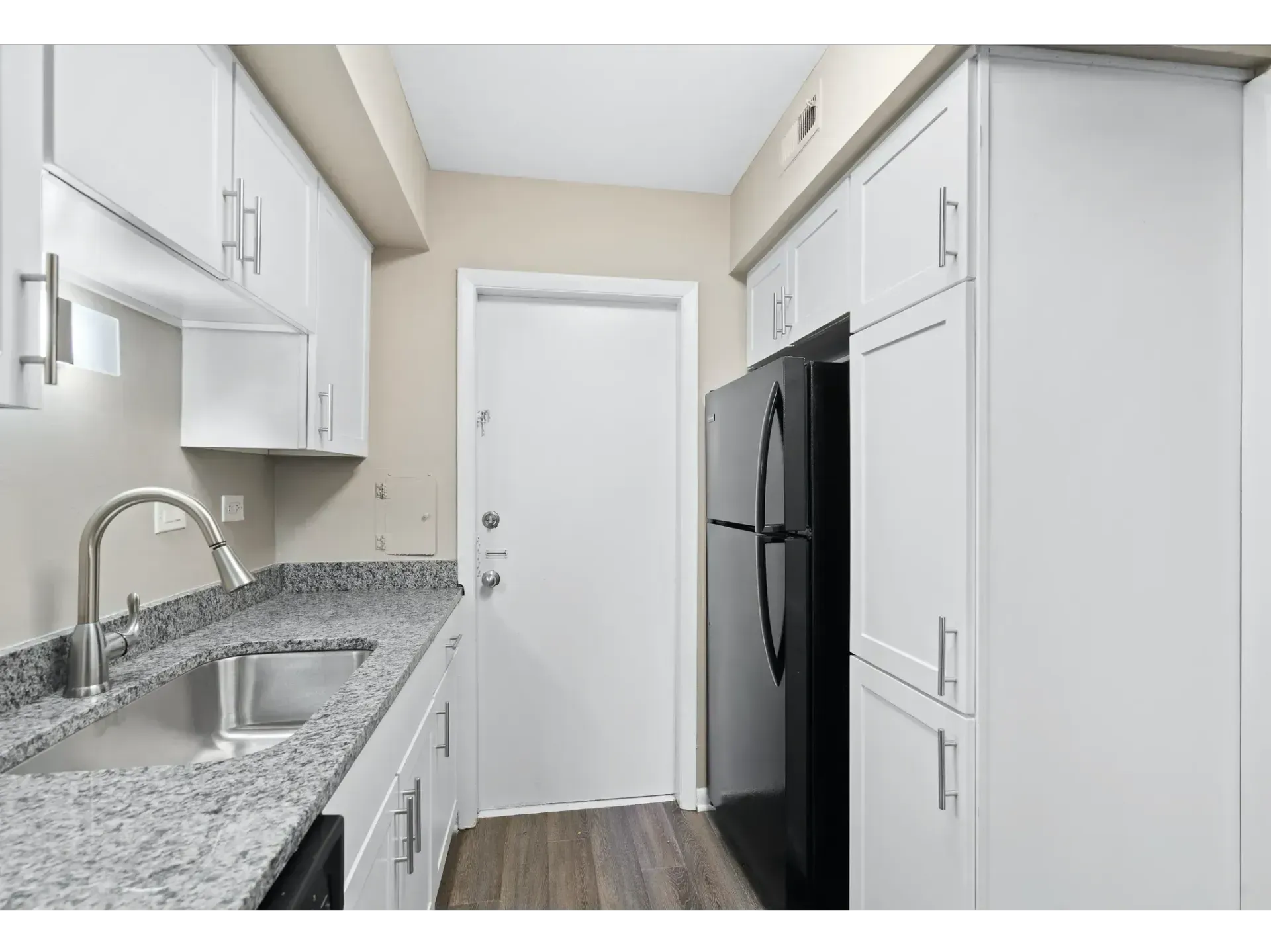 Narrow white kitchen with granite counters, stainless sink, black fridge, and closed door at the end
