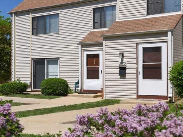 Two-story townhouse with a front walkway, white siding, brown roof, and purple flowers in the foreground