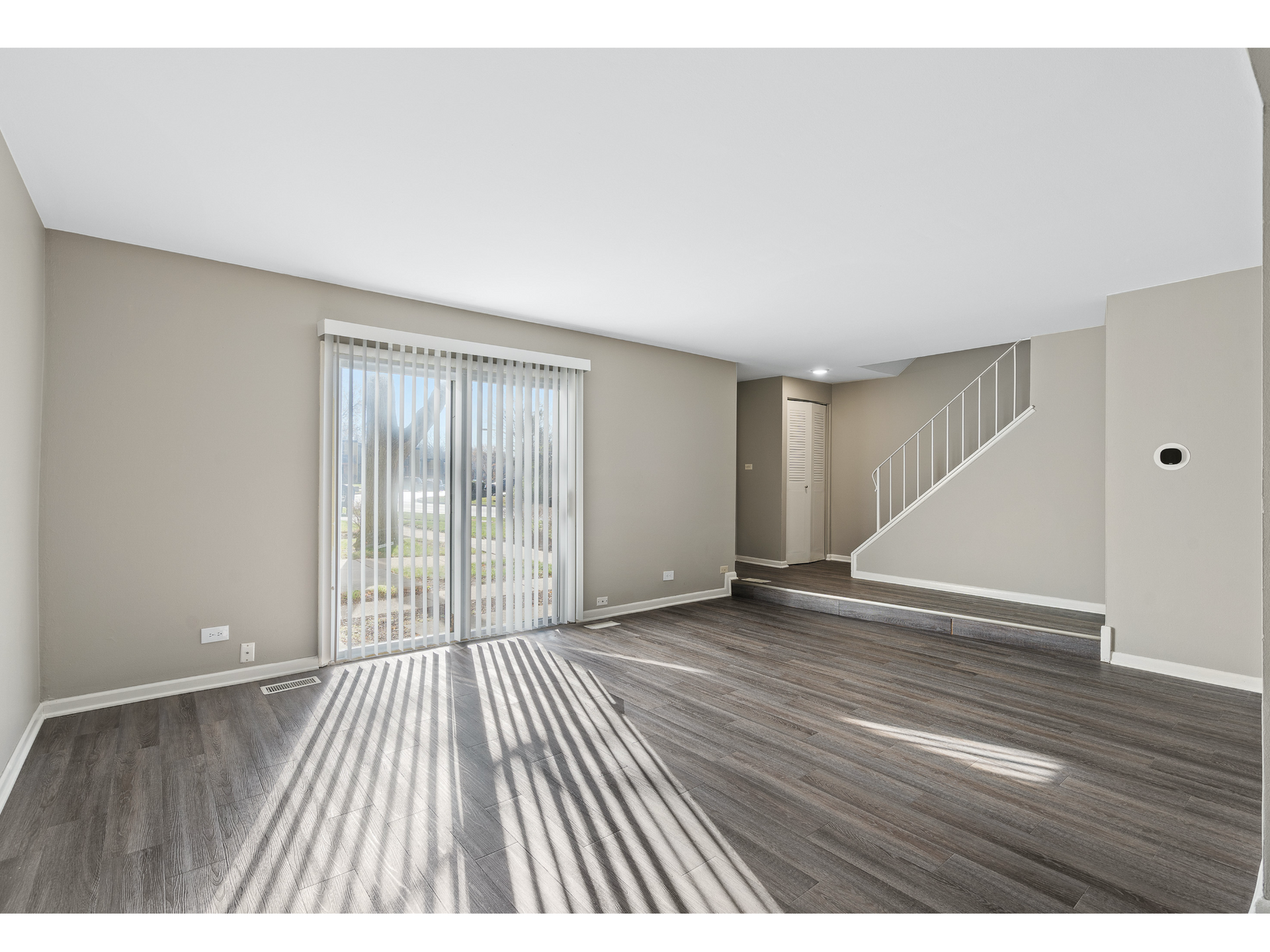 Empty living room with gray floors, beige walls, sliding glass door, and staircase with white railing.