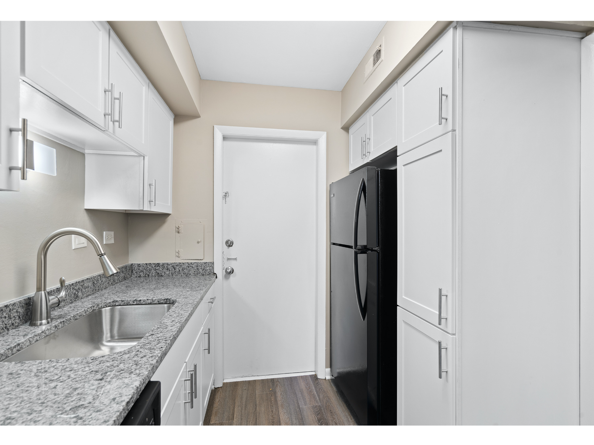 Narrow white kitchen with granite countertops, stainless sink, black refrigerator, and wood-look floor