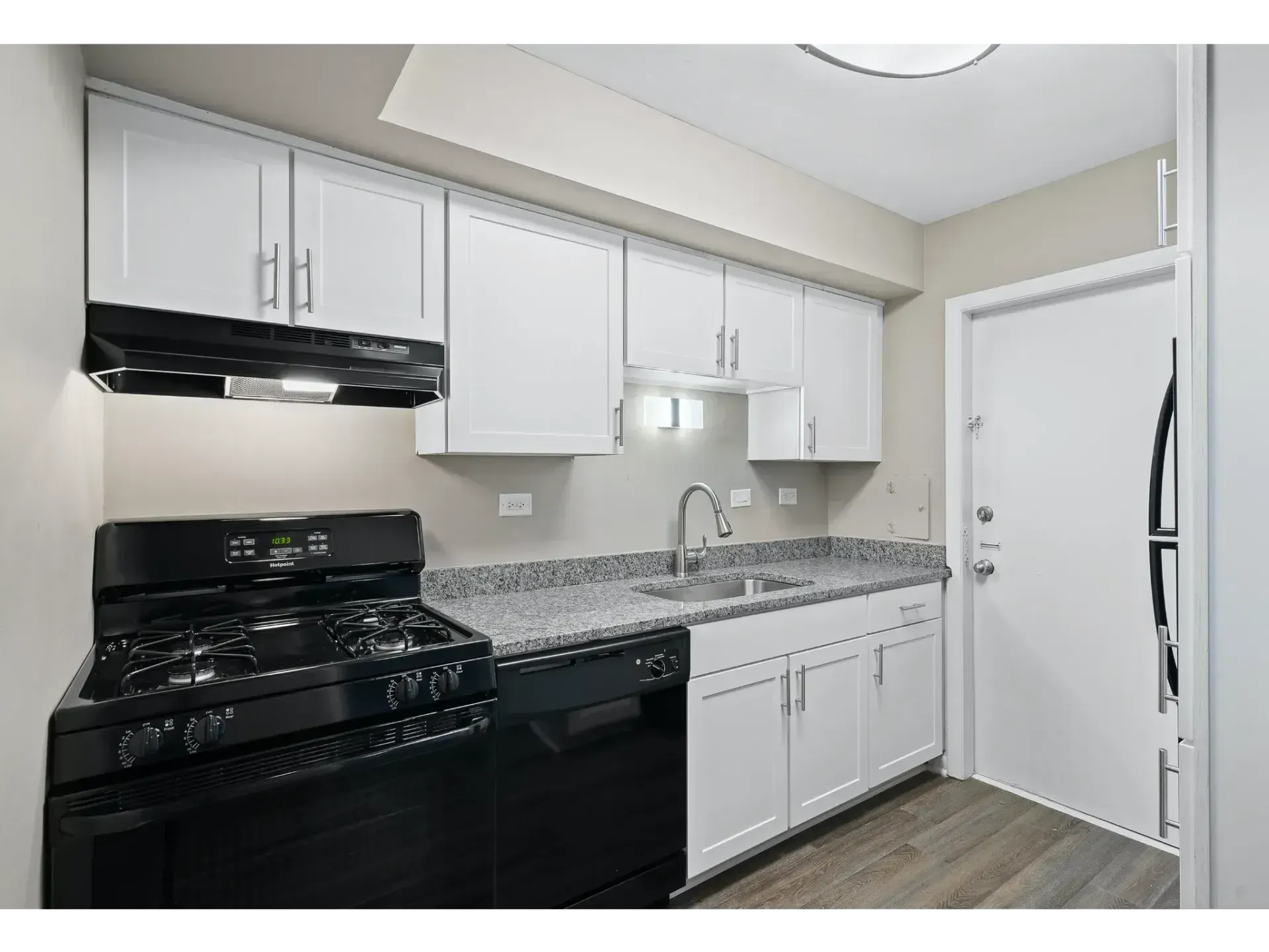 Small white kitchen with black stove, gray countertops, white cabinets, and a door by the sink.