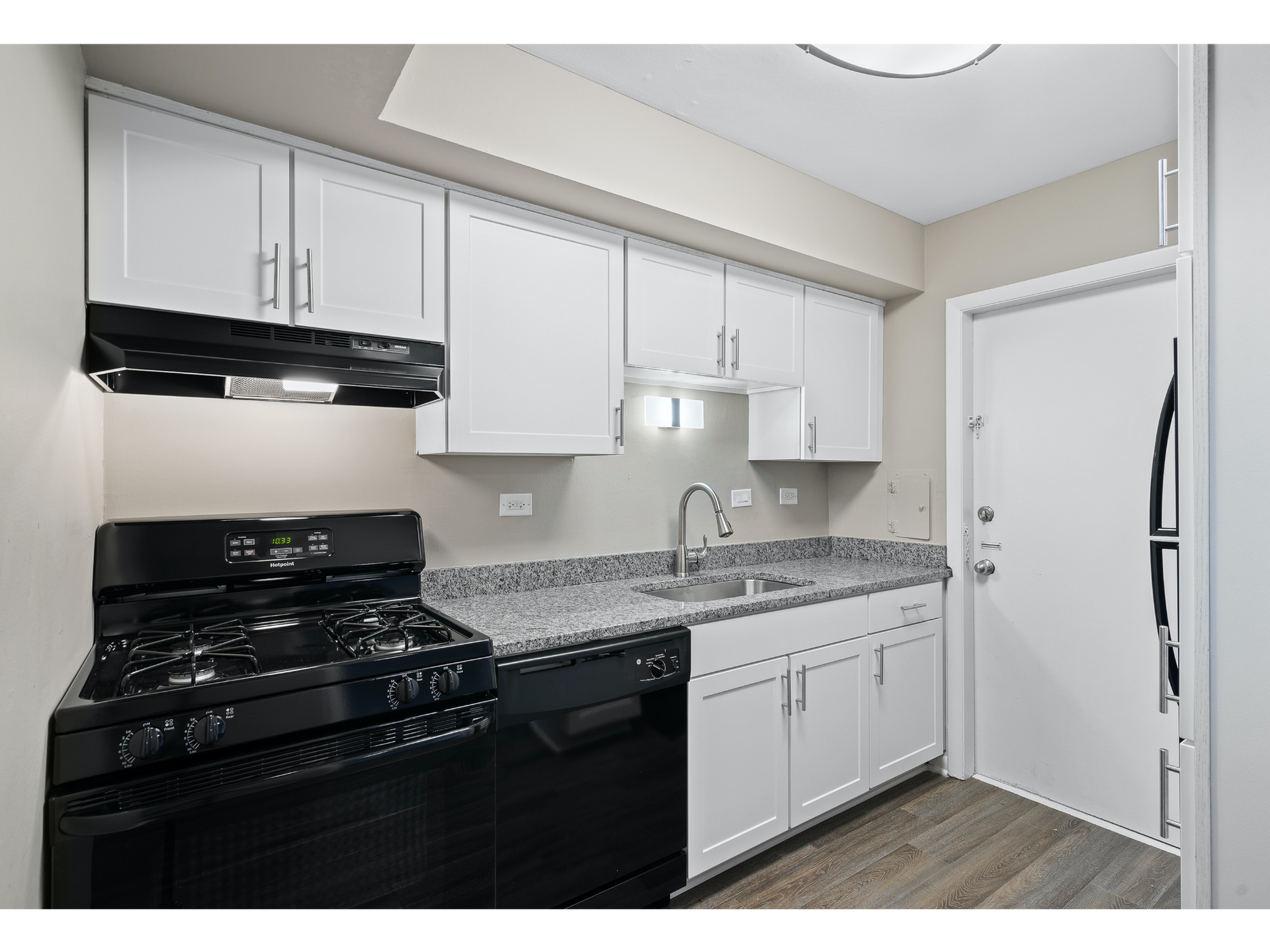 Small white kitchen with black stove, white cabinets, granite counters, and a door at right.