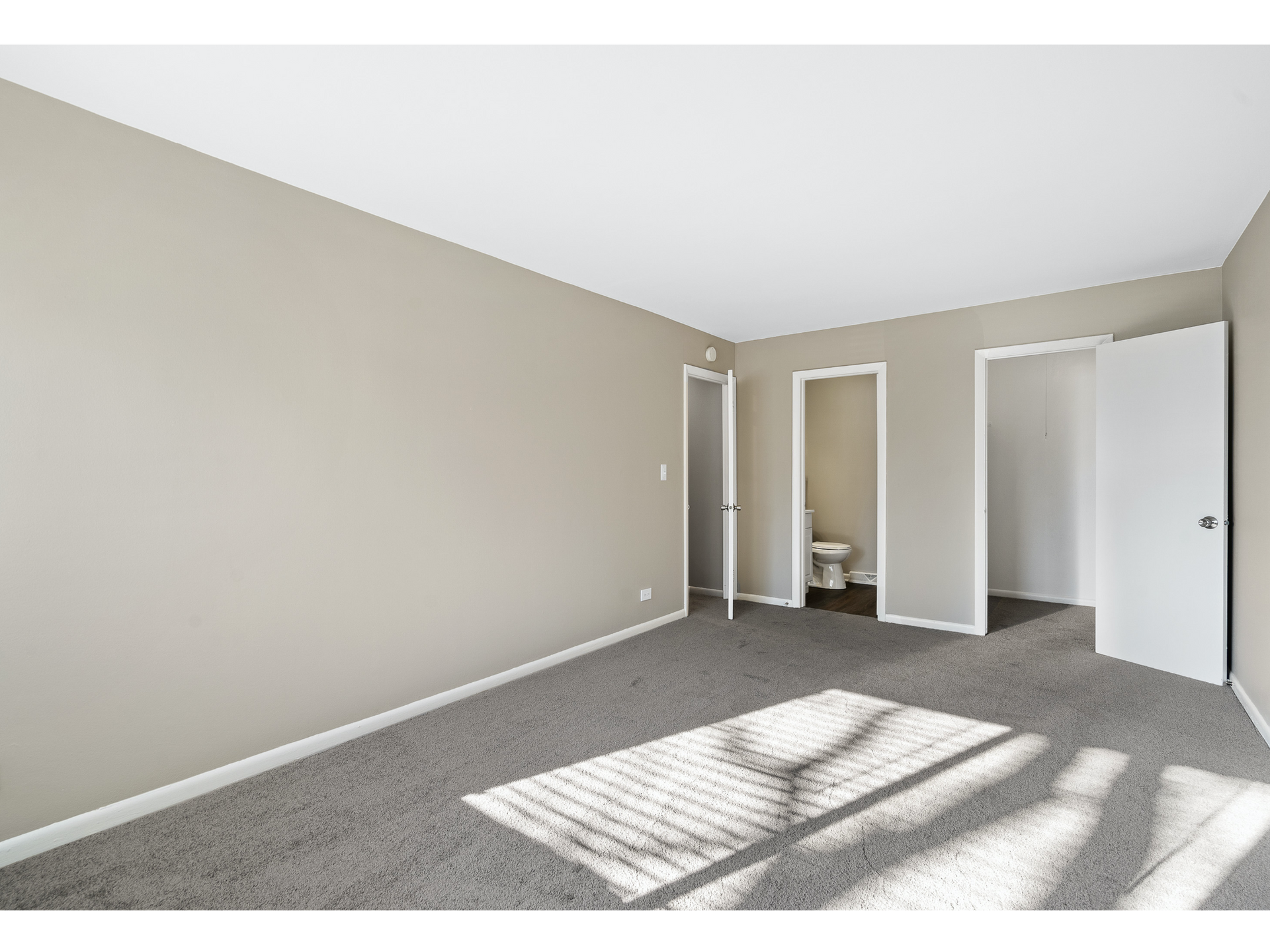Empty beige bedroom with gray carpet, sunlight on the floor, and open closet and doorway.