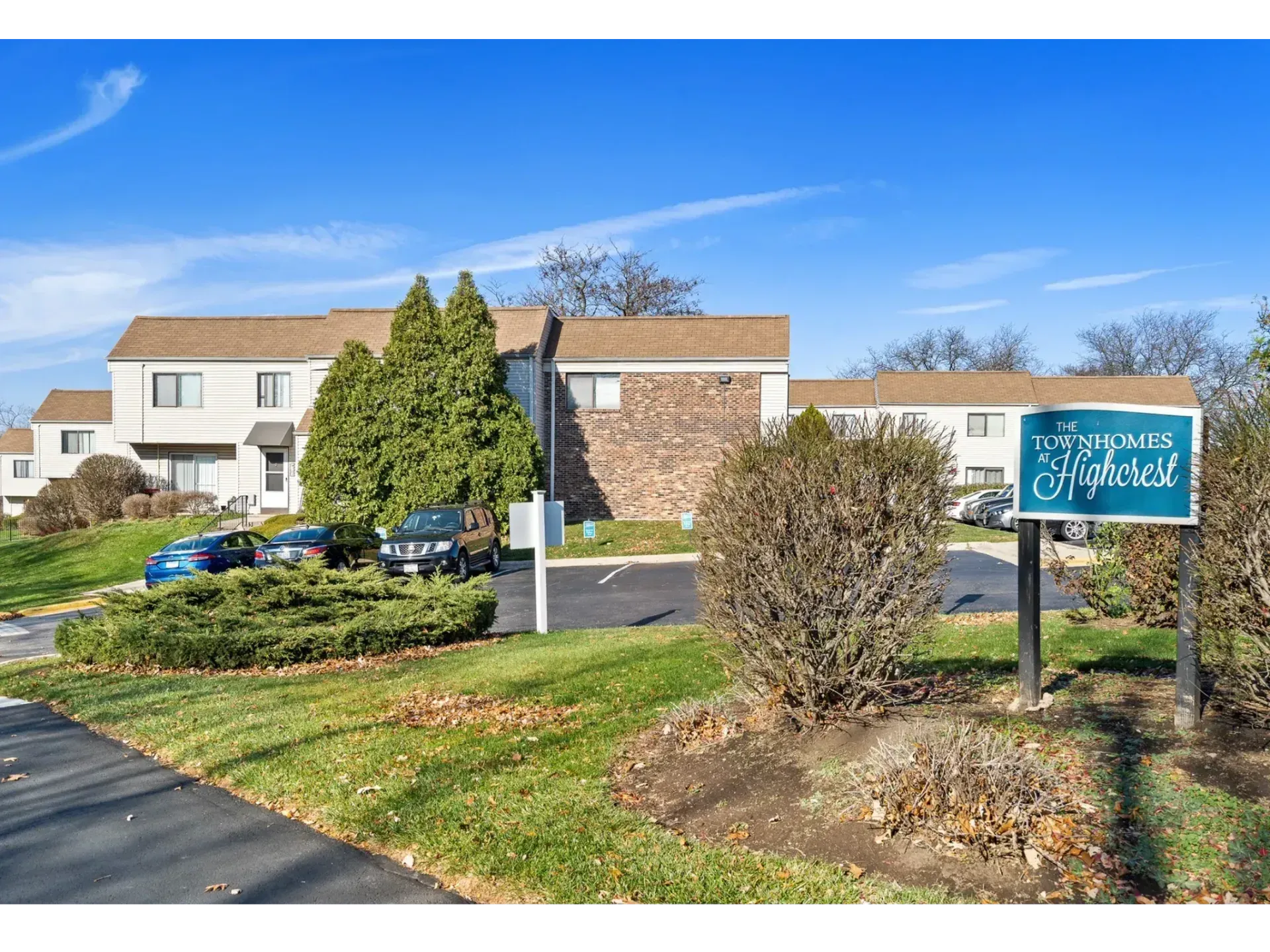 Apartment complex entrance sign with parked cars and shrubs on a sunny day