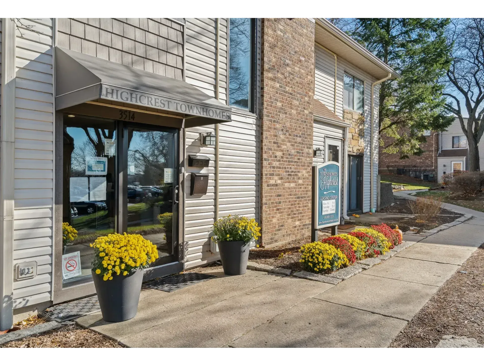 Storefront entrance with glass doors, potted yellow flowers, and a brick-and-siding exterior under a blue sky