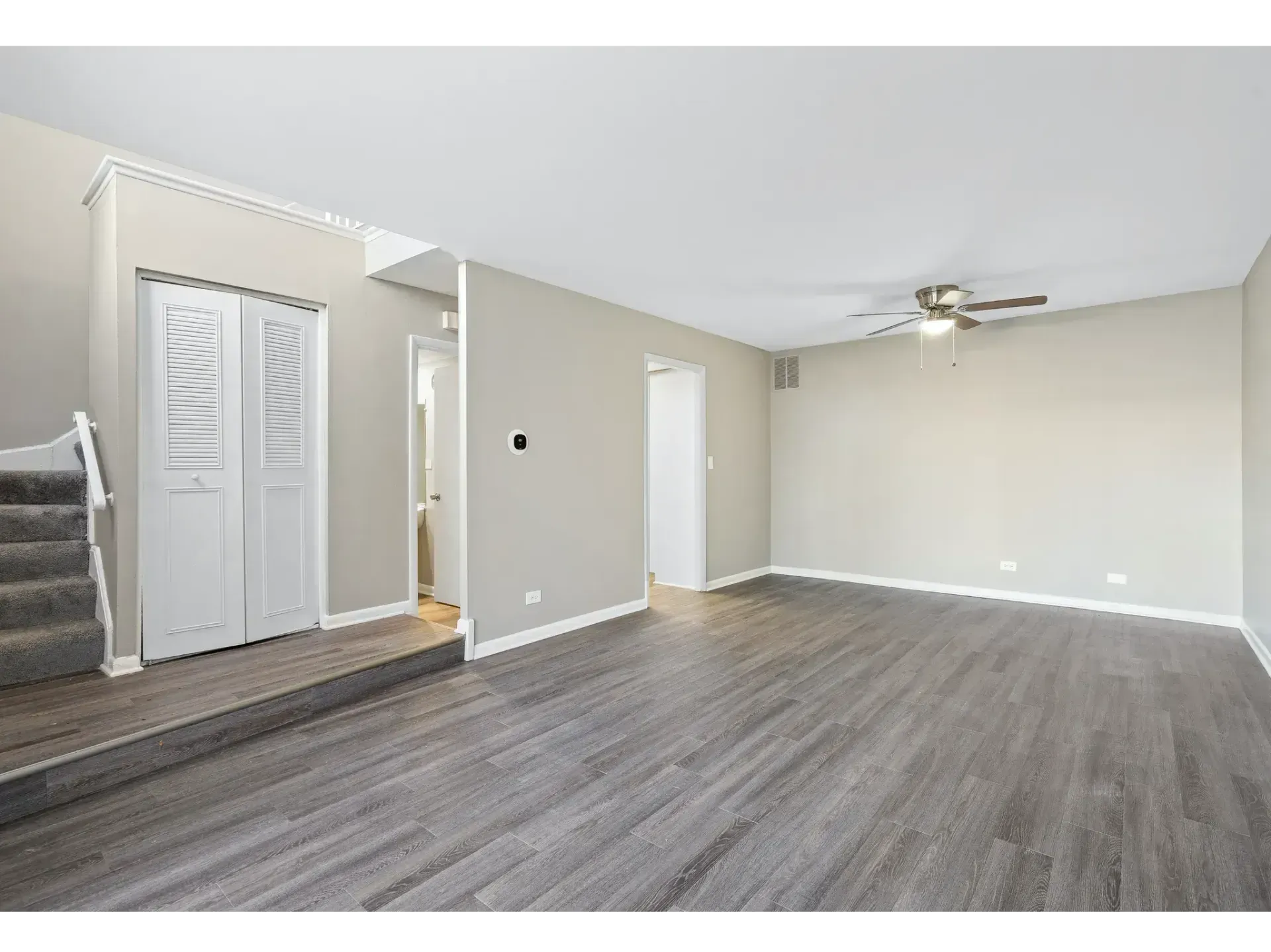 Empty living room with gray wood floors, beige walls, stairs, and a ceiling fan