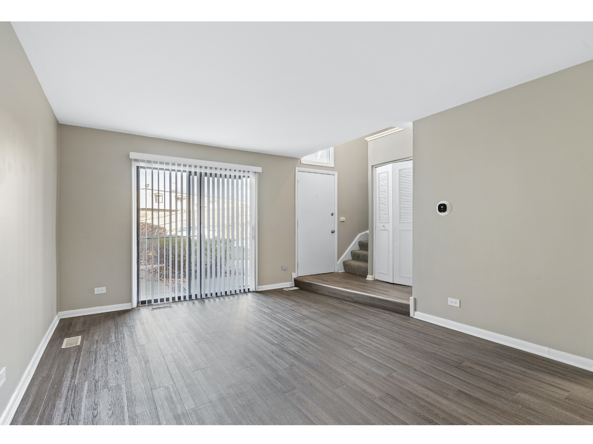 Empty beige living room with gray flooring and sliding glass doors, leading to a staircase and white closet doors