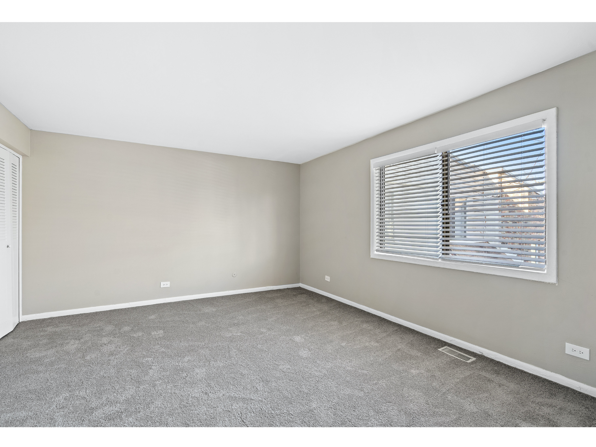Empty beige bedroom with gray carpet and a large window with blinds