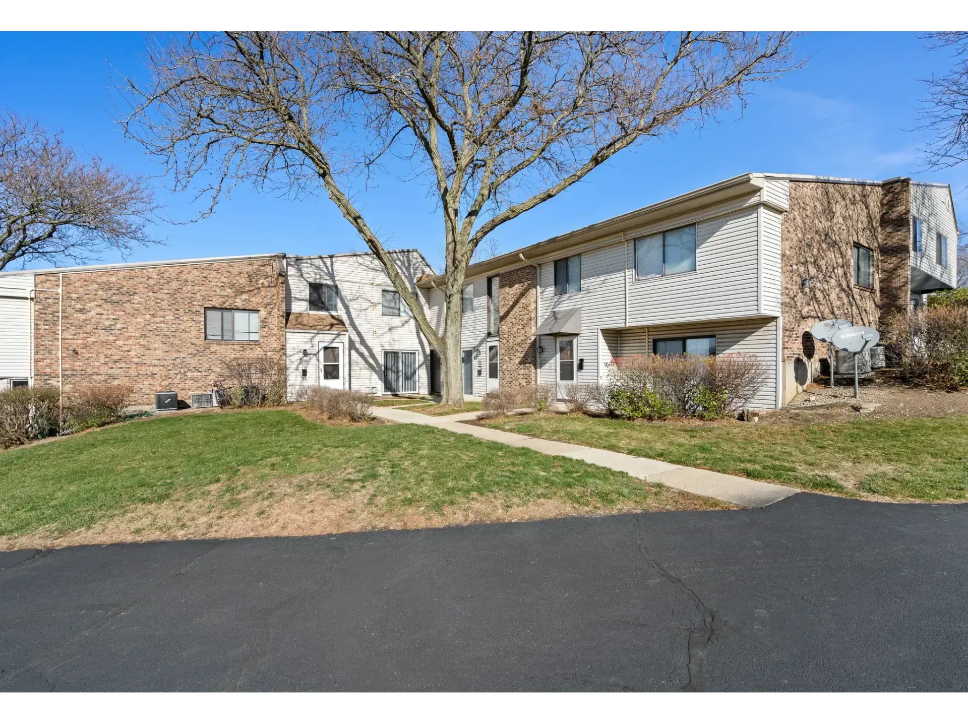 Two-story apartment building with brick and white siding, front walkway, and a leafless tree under a blue sky
