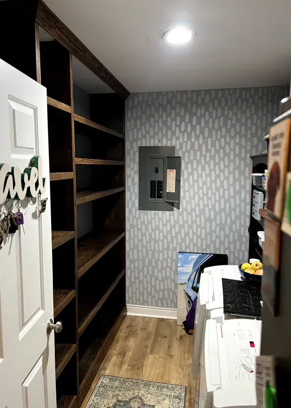 Pantry interior with built-in dark wooden shelves, white door, and grey patterned wallpaper.