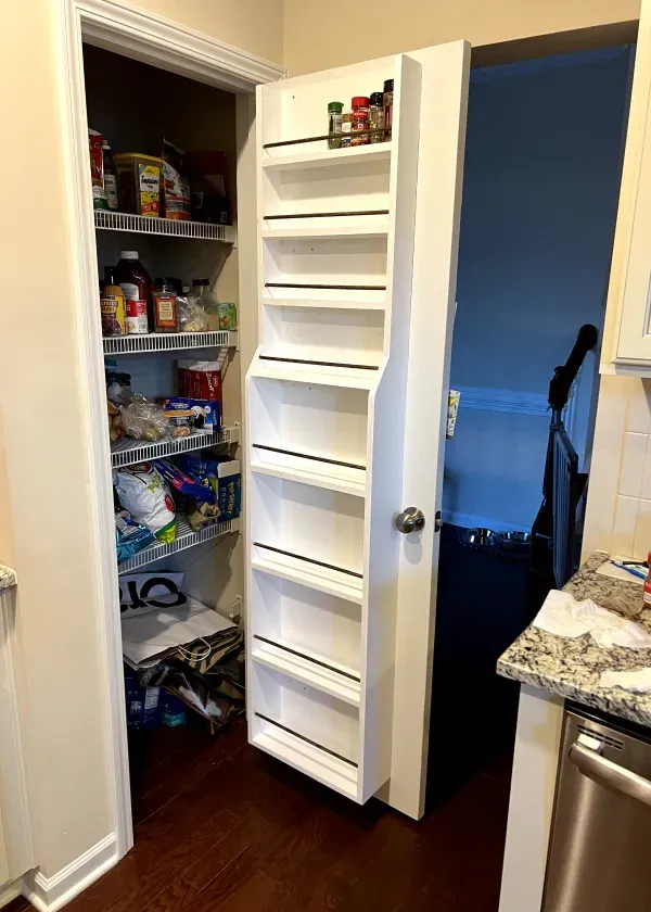 White pantry door with spice rack, opening into a cluttered pantry, kitchen setting.