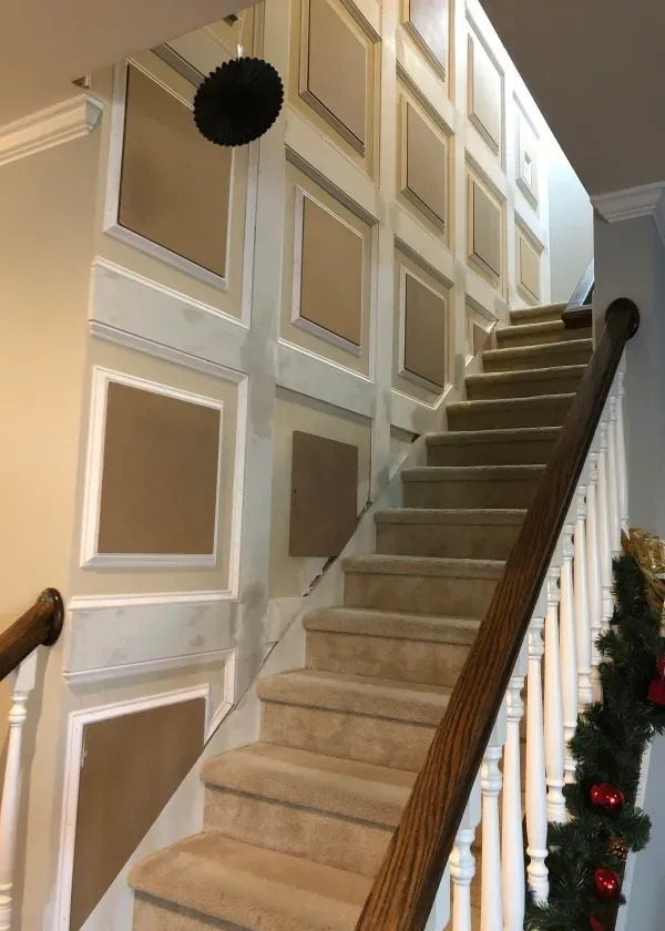 Staircase with framed wall panels. Tan carpet stairs with wooden handrails. Decorative Christmas garland on railing.