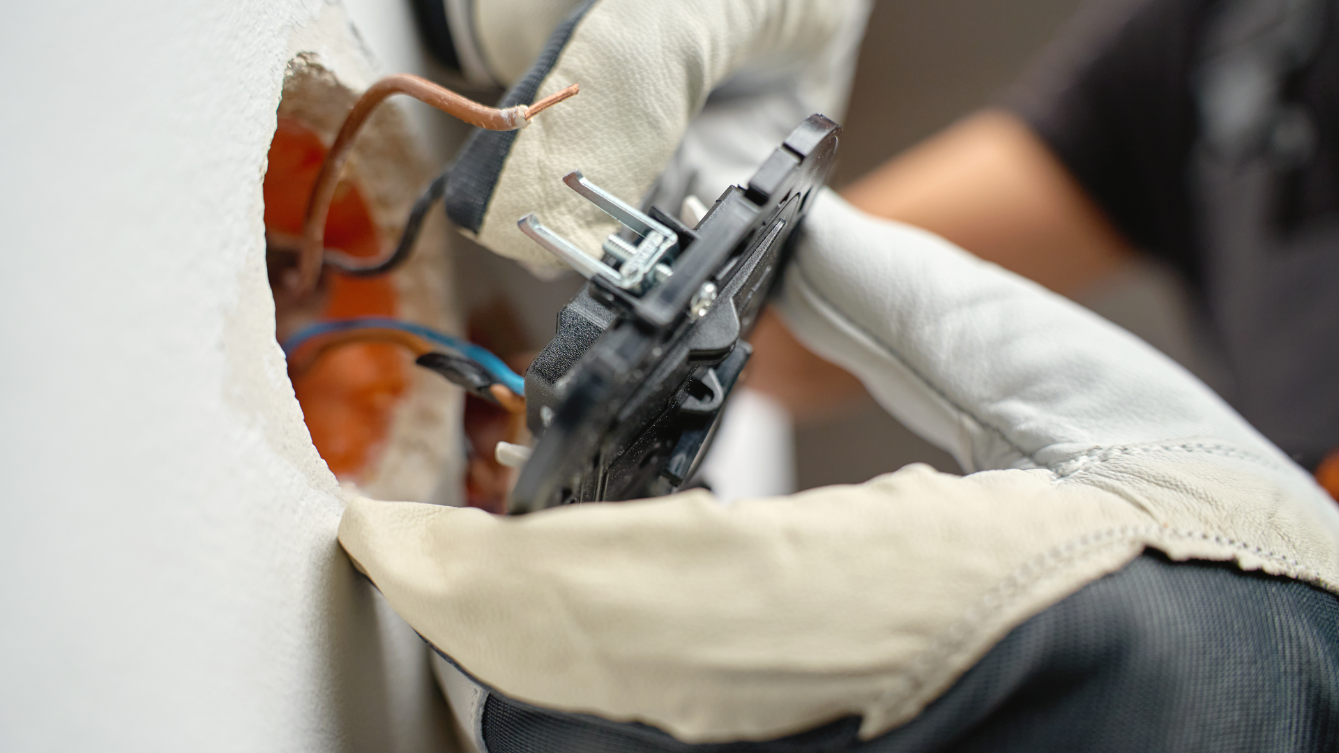 A person wearing protective gloves installs or repairs an electrical outlet on a white wall.