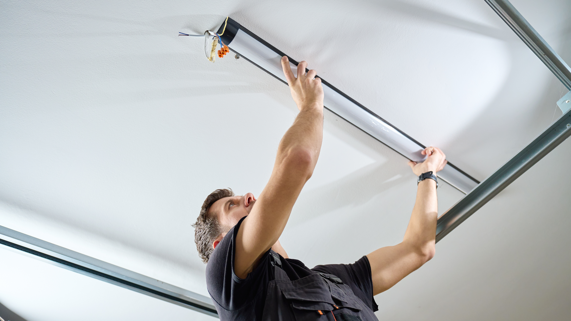 A person in a dark shirt installs a long, linear LED light fixture onto a white ceiling.