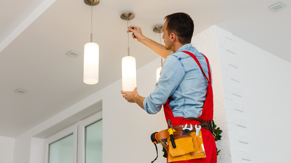 A person in red overalls and a tool belt adjusts a hanging pendant light in a white, modern interior.