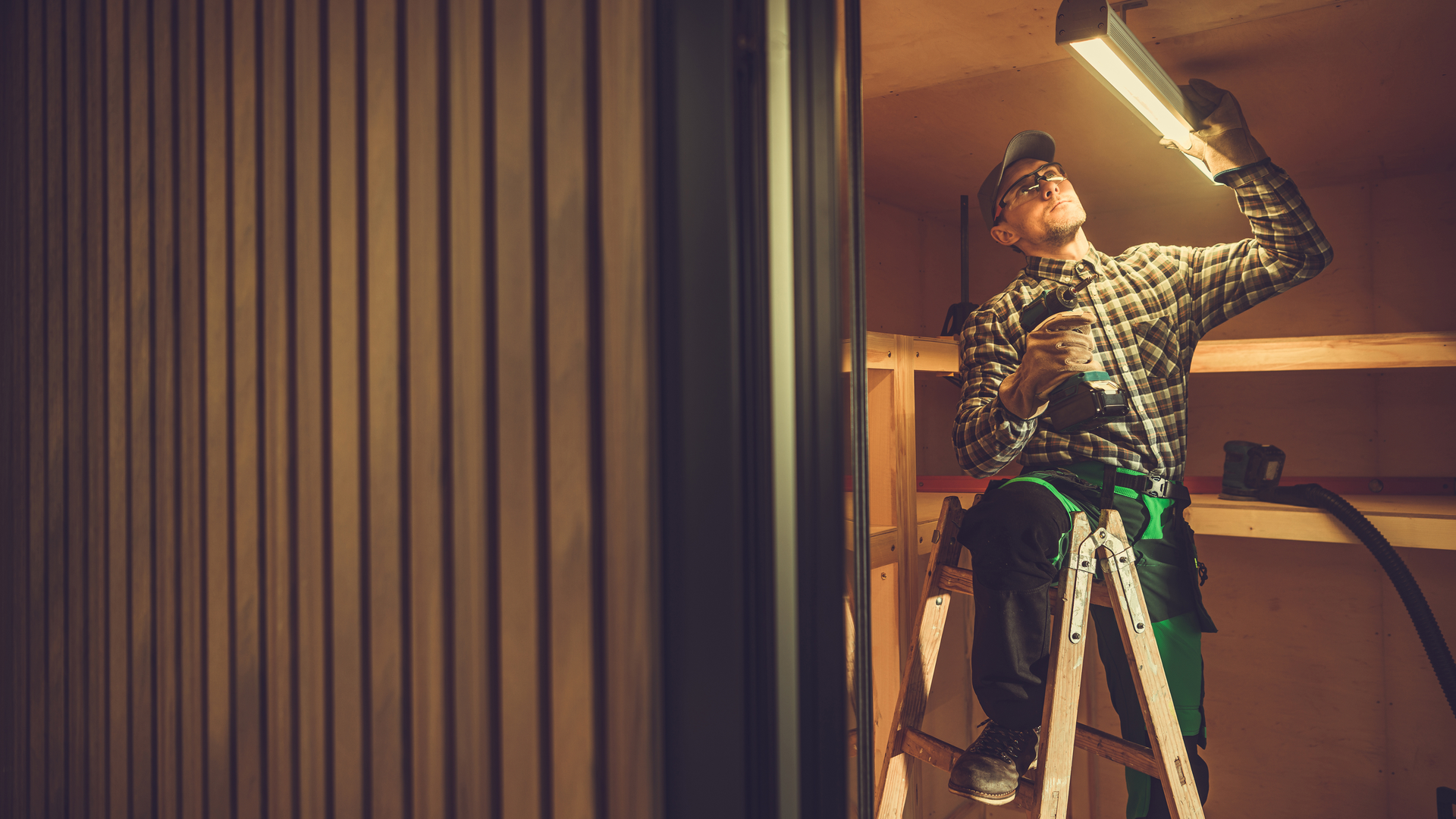 A person in workwear stands on a ladder, using a cordless drill to install a light fixture on a wooden ceiling.