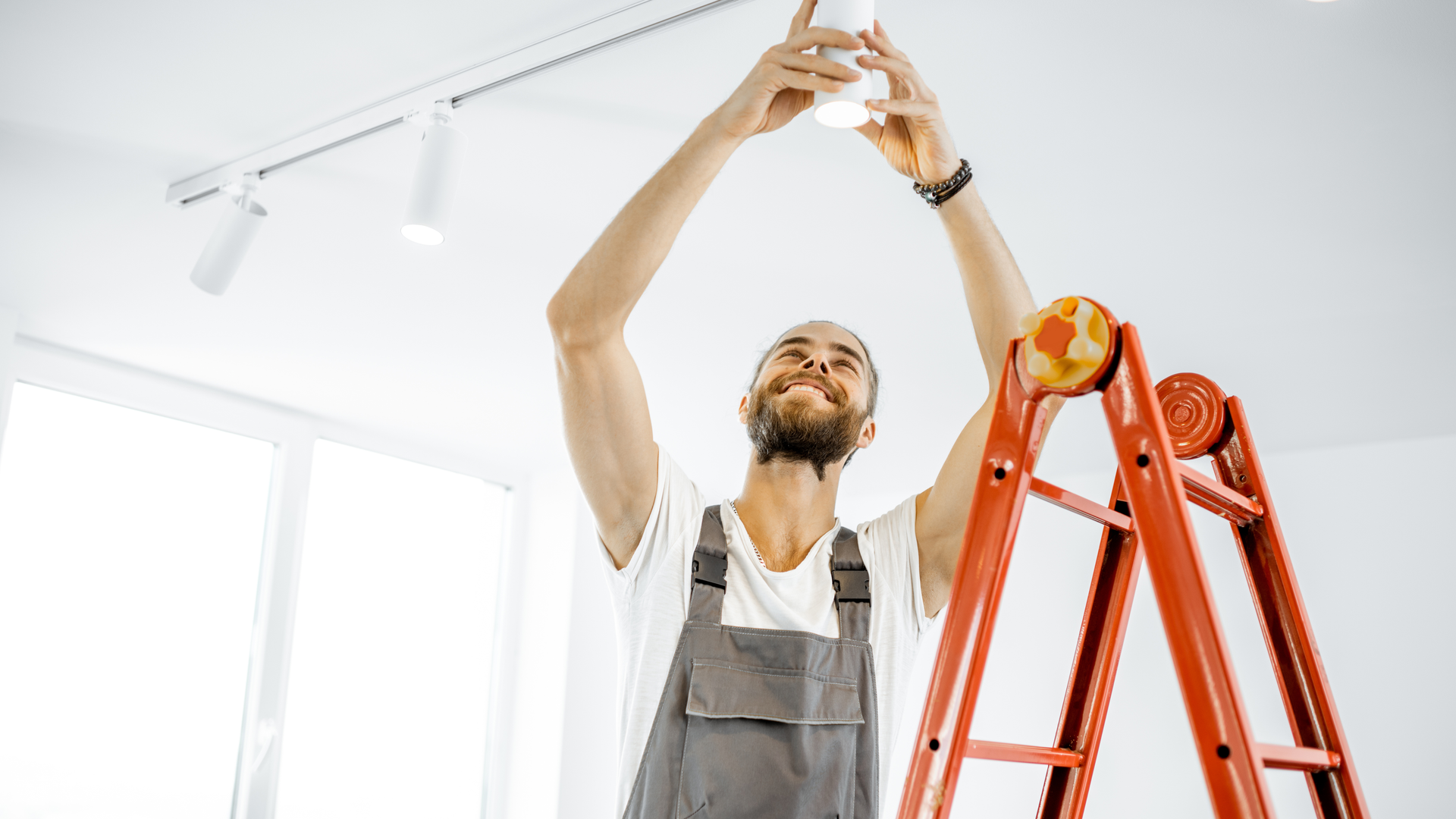 A smiling person in work overalls standing on a red ladder while installing a ceiling light fixture in a bright room.
