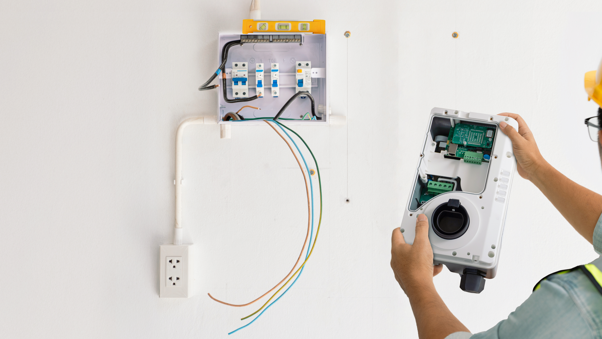 An electrician holding a detached device part, preparing to connect it to wall-mounted circuit breakers and wiring.