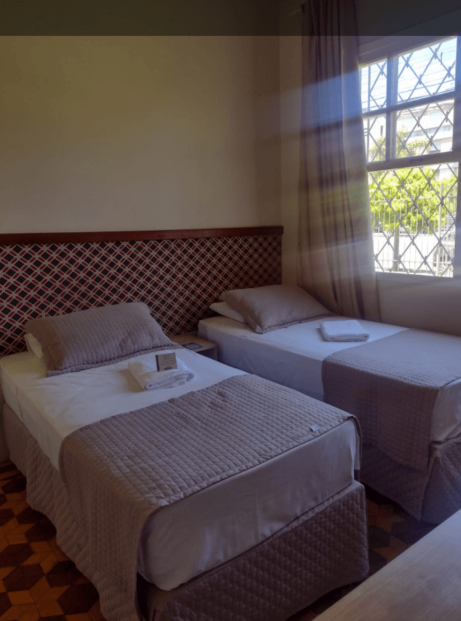 Um quarto de hotel com duas camas de solteiro com lençóis brancos, peseiras e travesseiros bege, em frente a uma cabeceira estampada.