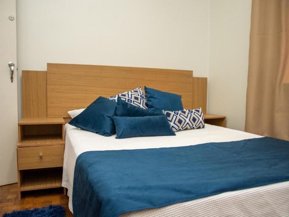 Uma cama bem arrumada com cabeceira de madeira clara, travesseiros azuis e uma peseira azul em um quarto simples.