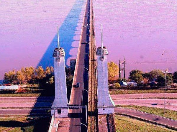 Vista aérea de uma ponte com altos pilares brancos ladeando uma estrada e trilhos de trem que se estendem sobre um rio.