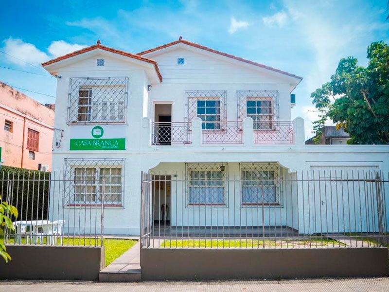 Um prédio branco de dois andares com uma placa verde e grades de metal nas janelas, atrás de um gramado frontal cercado.