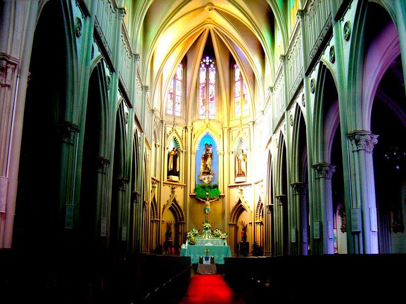 Vista interior de uma igreja em estilo gótico com tetos altos abobadados, colunas, um altar central e um tapete vermelho no corredor central.
