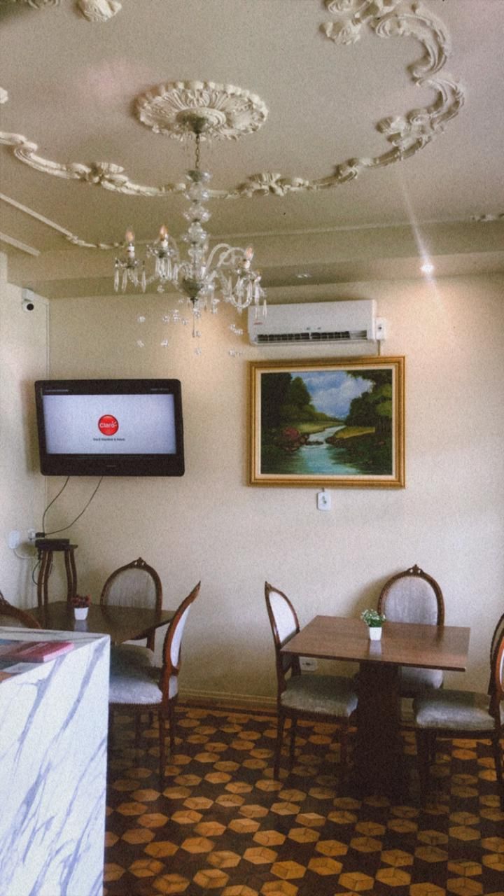 Uma sala de jantar com mesas quadradas, cadeiras, uma TV fixada na parede, um quadro com uma paisagem e uma luminária de teto ornamentada.