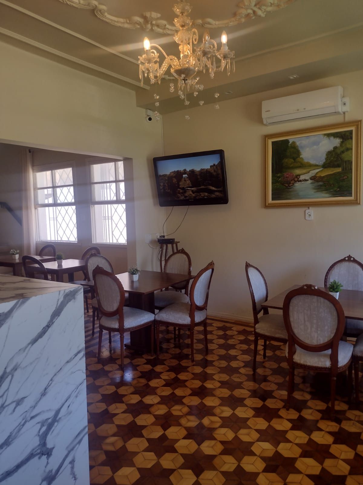 Uma sala de jantar com mesas redondas e cadeiras cor creme, um lustre de cristal, uma TV fixada na parede e uma pintura de paisagem.