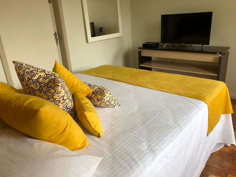 Uma cama com roupa de cama branca, almofadas decorativas amarelas e um tapete amarelo num quarto com uma televisão sobre um suporte.