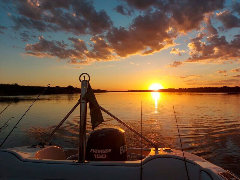Pôr do sol sobre águas calmas, visto da parte traseira de um barco equipado com varas de pesca e um motor de popa.