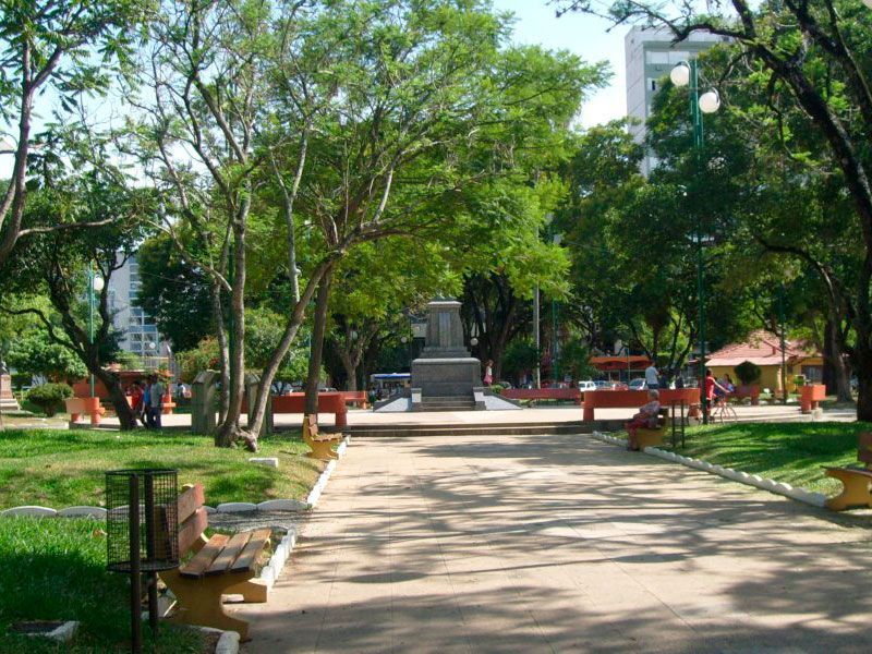 Uma praça ensolarada e arborizada da cidade, com um monumento central, caminhos pavimentados e vários bancos de madeira.