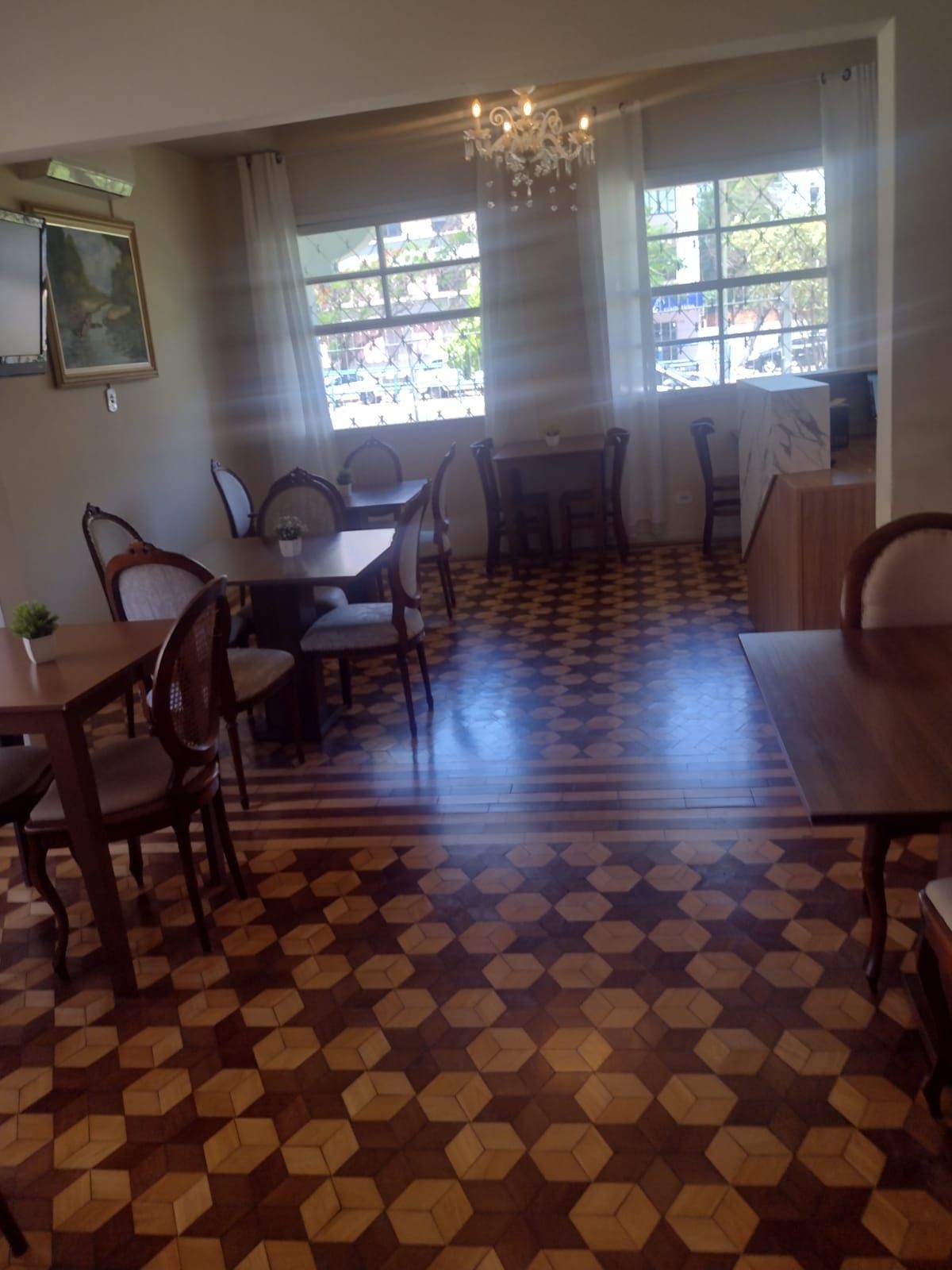 Sala de jantar de um restaurante com mesas e cadeiras de madeira, e um piso com um padrão geométrico distinto sob um lustre.