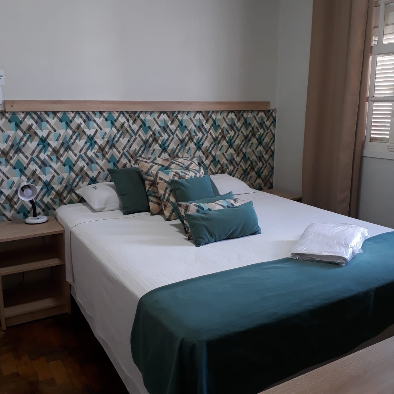 Um quarto com uma cama queen-size com cabeceira geométrica azul-petróleo, lençóis brancos, detalhes em azul-petróleo e uma mesa de cabeceira de madeira.