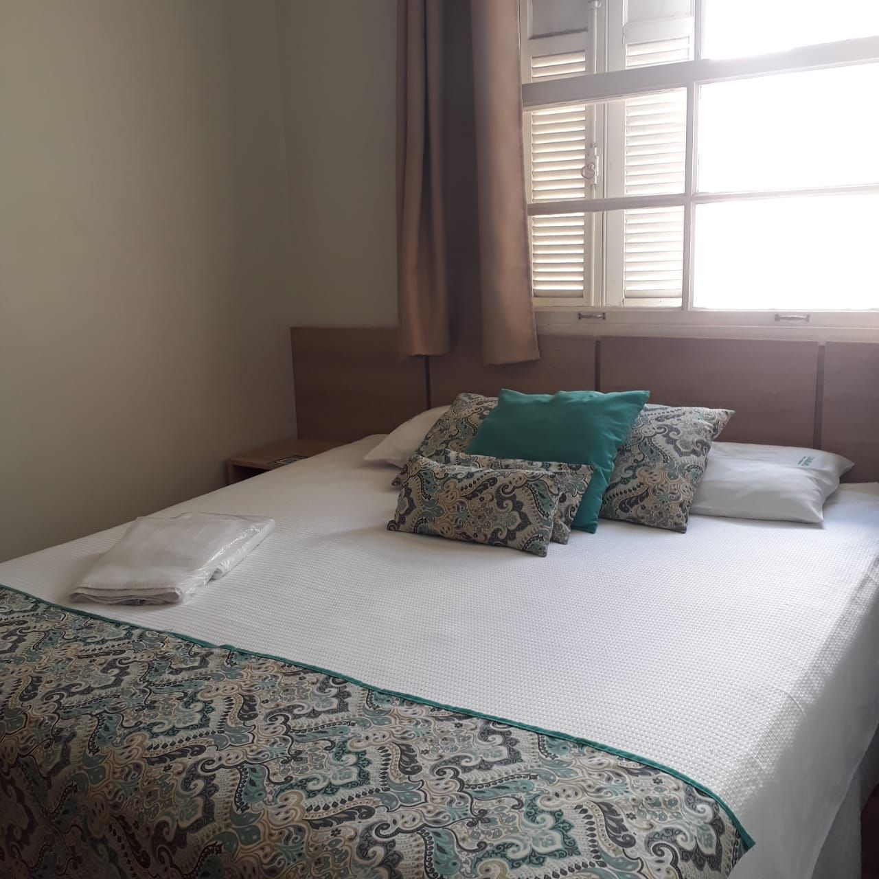 Uma cama com roupa de cama branca, um caminho de mesa decorativo em tons de azul-petróleo com estampa e almofadas decorativas combinando em frente a uma janela.