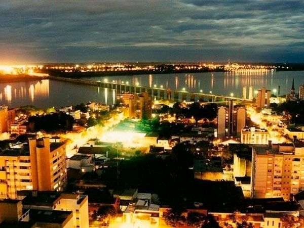 Vista aérea de uma cidade à noite, com prédios iluminados, postes de luz e uma ponte cruzando um rio escuro.