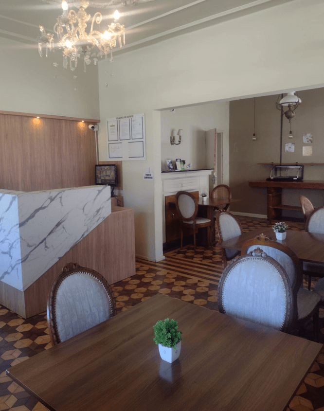 Área de jantar de um restaurante vazia, com mesas marrons, cadeiras estofadas, balcão de atendimento com estampa de mármore e piso de azulejo.