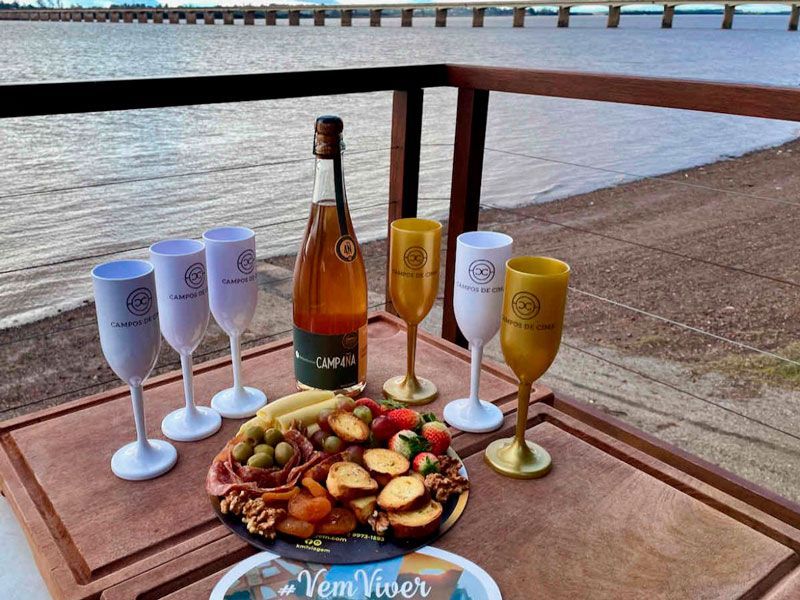 Uma mesa de madeira com uma garrafa de vinho, seis copos variados e uma tábua de charcutaria com vista para um corpo d'água e uma ponte.