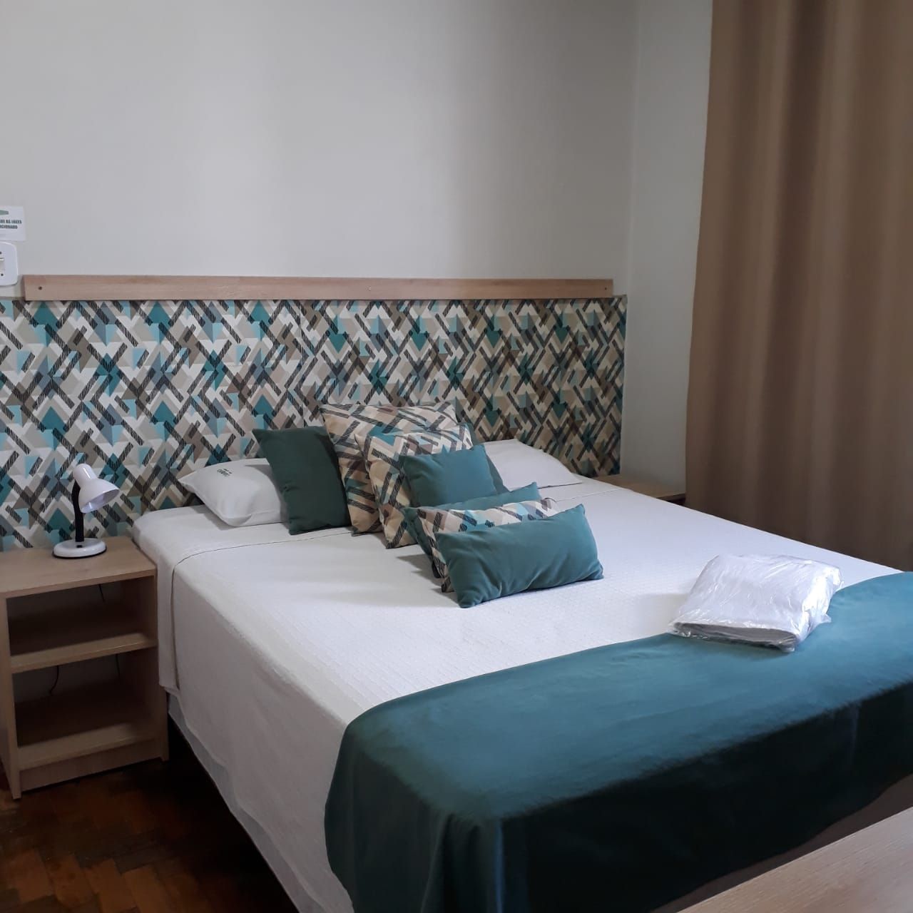 Um quarto de hotel bem arrumado apresenta uma cama de casal com uma peseira azul-petróleo, travesseiros combinando e uma cabeceira com estampa geométrica.