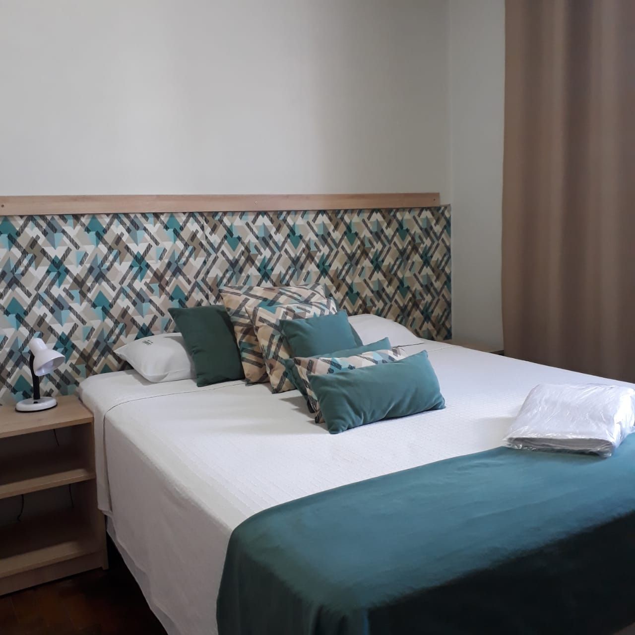 Uma cama arrumada com travesseiros verde-azulados e um caminho de cama combinando, em frente a uma cabeceira com papel de parede geométrico estampado, em um quarto.