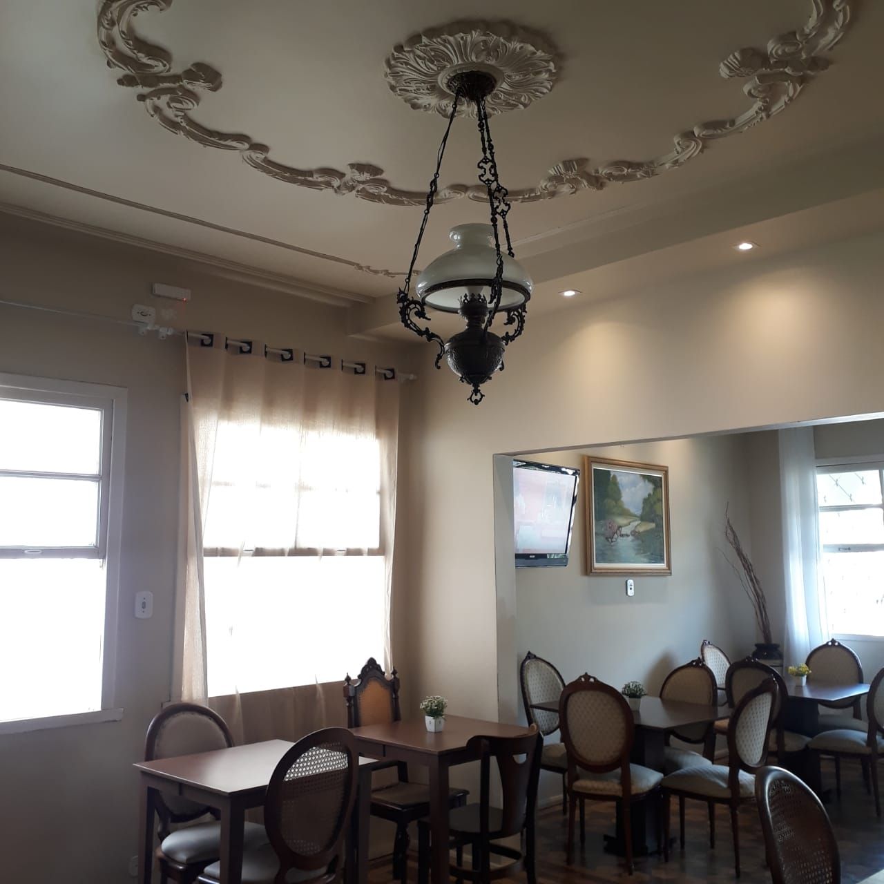 Uma área de jantar interna com mesas e cadeiras, um candeeiro suspenso em estilo vintage e um medalhão decorativo no teto.