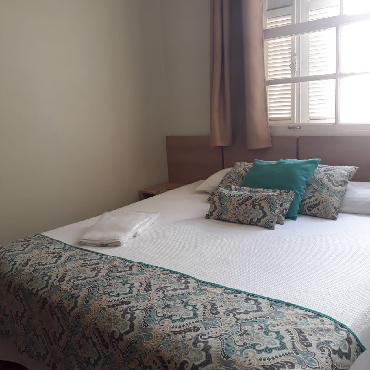 Uma cama bem arrumada com um edredom branco e almofadas decorativas em tons de verde-azulado e estampadas, situada ao lado de uma janela.