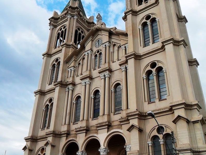 Foto tirada em ângulo baixo de uma catedral neoclássica de pedra bege com duas torres altas sob um céu azul nublado.