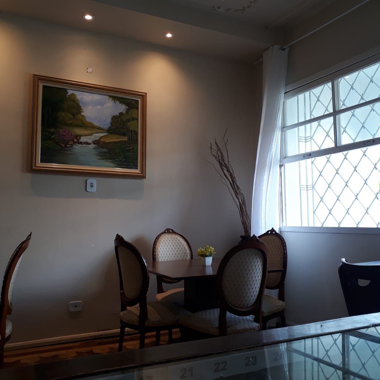 Um canto de jantar com uma pequena mesa, quatro cadeiras, um quadro com uma paisagem e um vaso com galhos secos perto de uma janela.