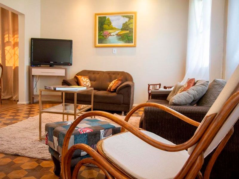 Uma sala de estar aconchegante com um sofá marrom, um pufe estampado, uma poltrona de madeira, uma TV em um suporte e obras de arte na parede.