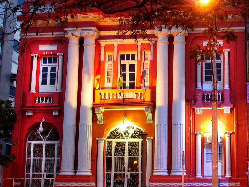 Um edifício histórico neoclássico de dois andares, pintado de um vermelho vibrante, com pilares brancos e portas em arco.