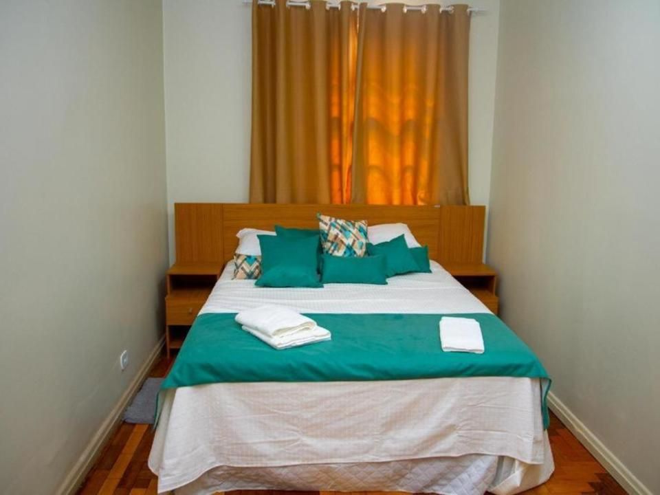 Uma cama bem arrumada com travesseiros verde-azulados e uma peseira combinando, posicionada contra uma cabeceira de madeira em um quarto mobiliado de forma simples.